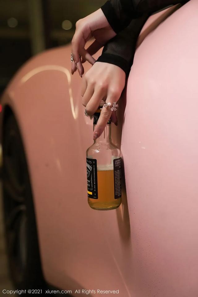 A woman holding a bottle of whiskey in front of a pink car.