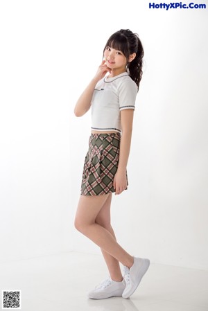 A woman in a white shirt and plaid skirt leaning against a wall.